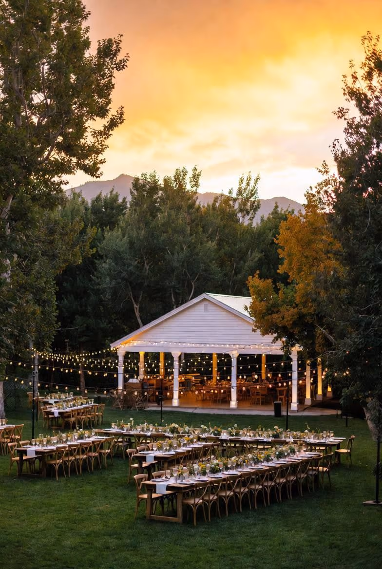 Outdoor wedding reception at a park pavilion at sunset with string lights and long farm tables